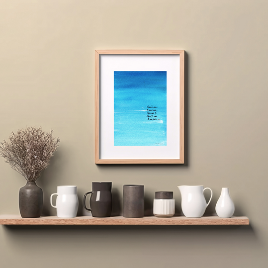 Framed abstract painting in blue with expressive brushstrokes and handwritten words Here I Am with a dot of red paint, spiritual modern art print, hanging on wall above a wooden counter top.