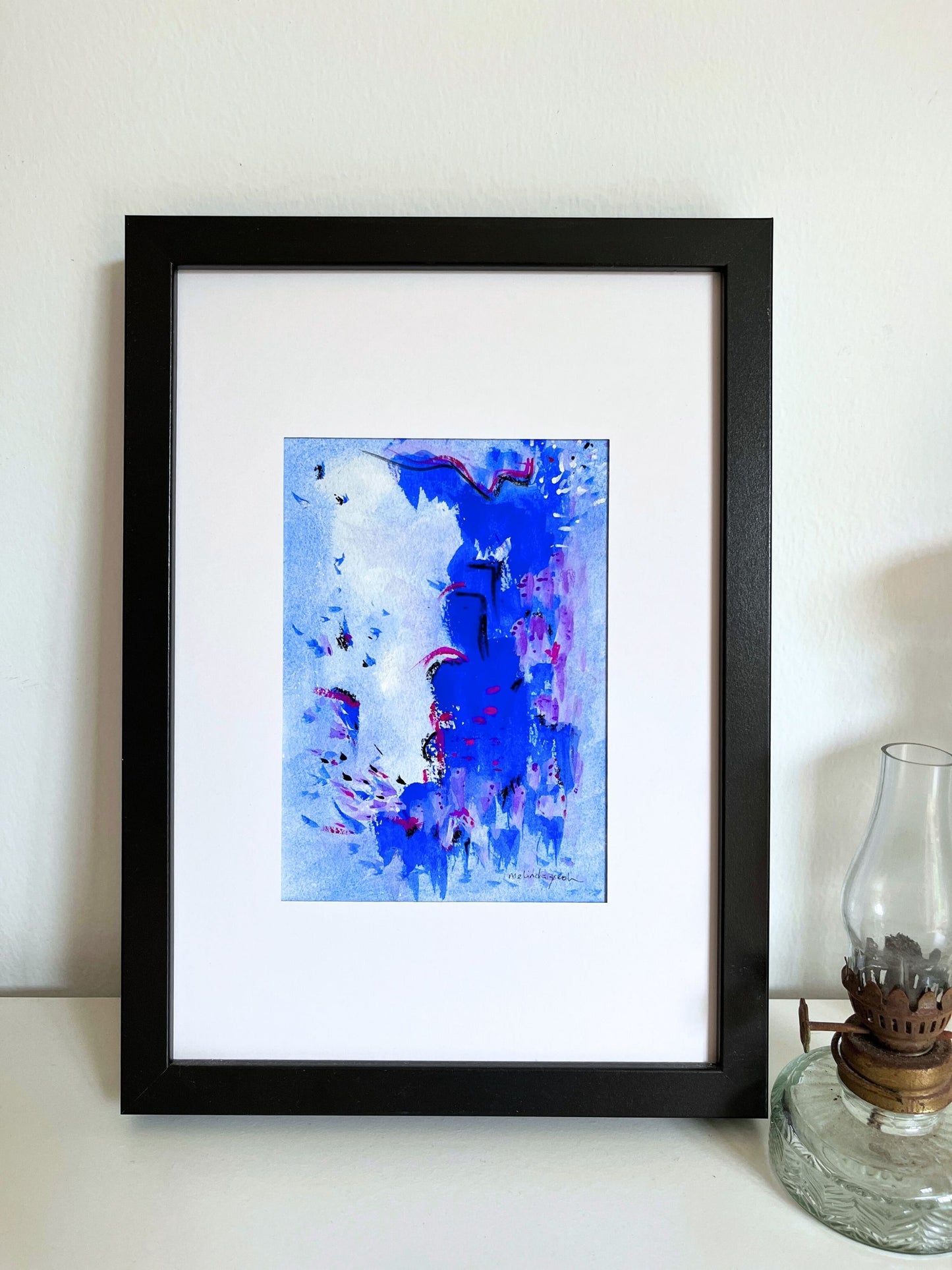 Framed abstract artwork with blue and purple hues on a white wall.