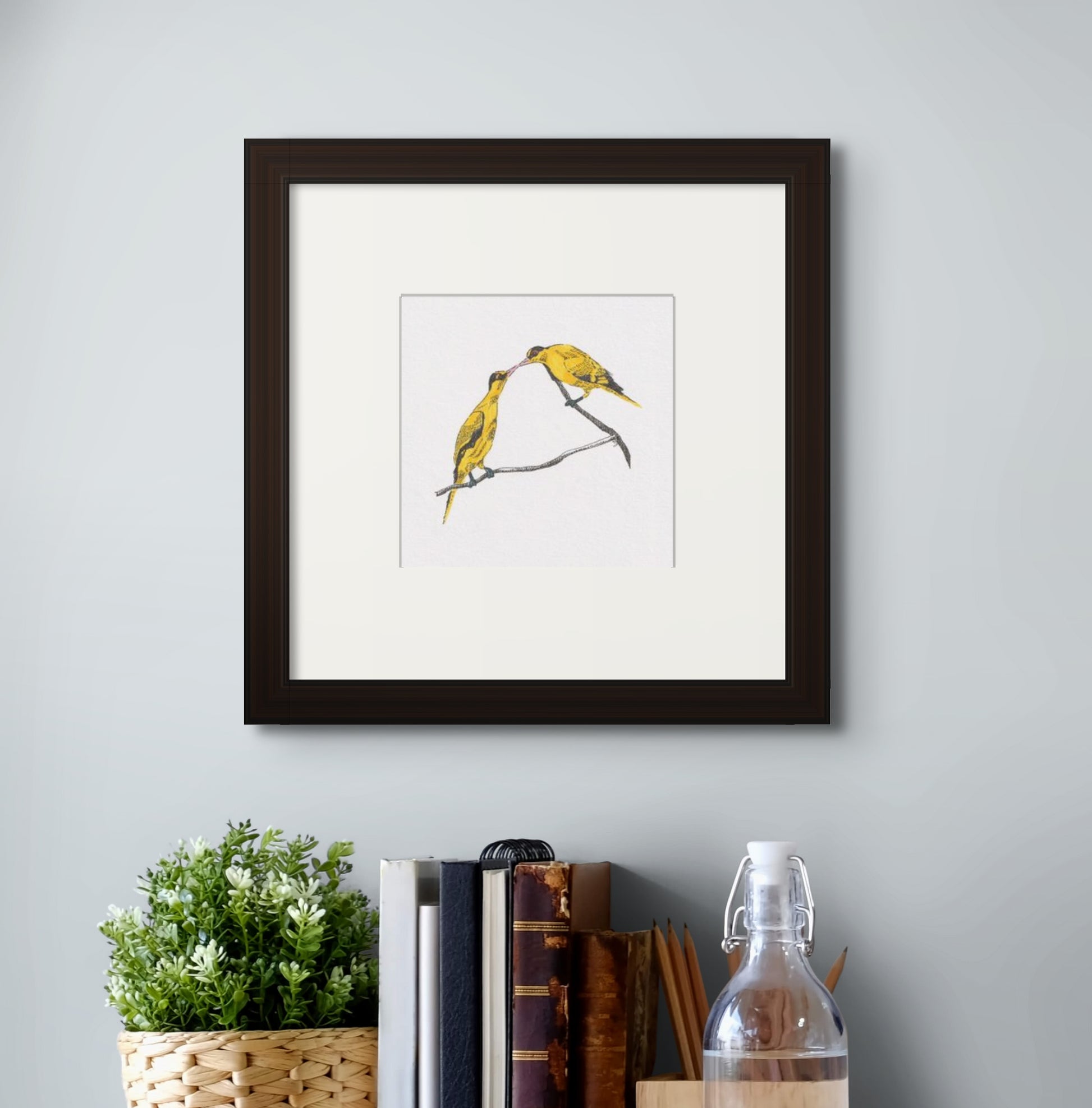 6” x 6” giclée art print of line drawing with coloured brush pen drawing of a pair of yellow oriole birds, with their beaks touching. In a black frame and cream colour mat. Hung above a work desk table top.