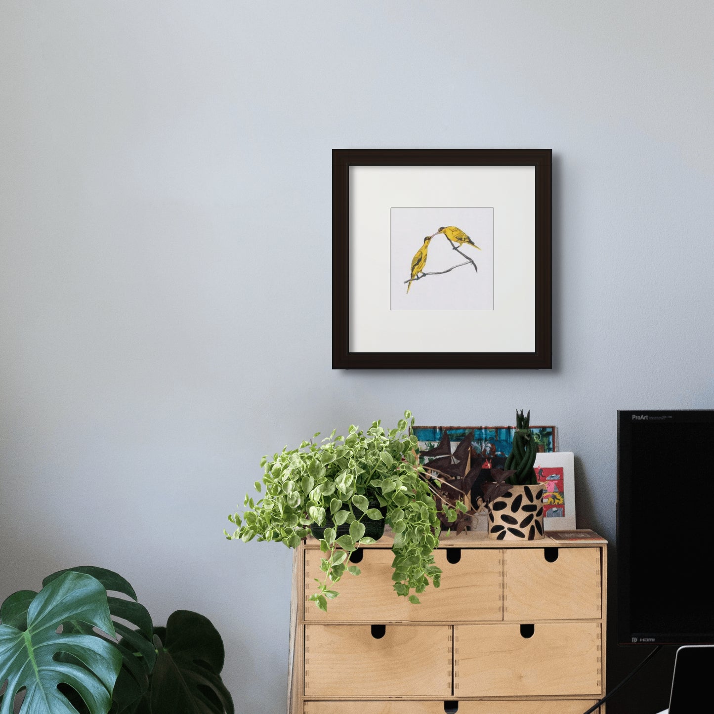 6” x 6” giclée art print with black frame and cream colour mat, in a home office setting. Line drawing with coloured brush pen drawing of a pair of yellow oriole birds, with their beaks touching. In a black frame and cream colour mat.