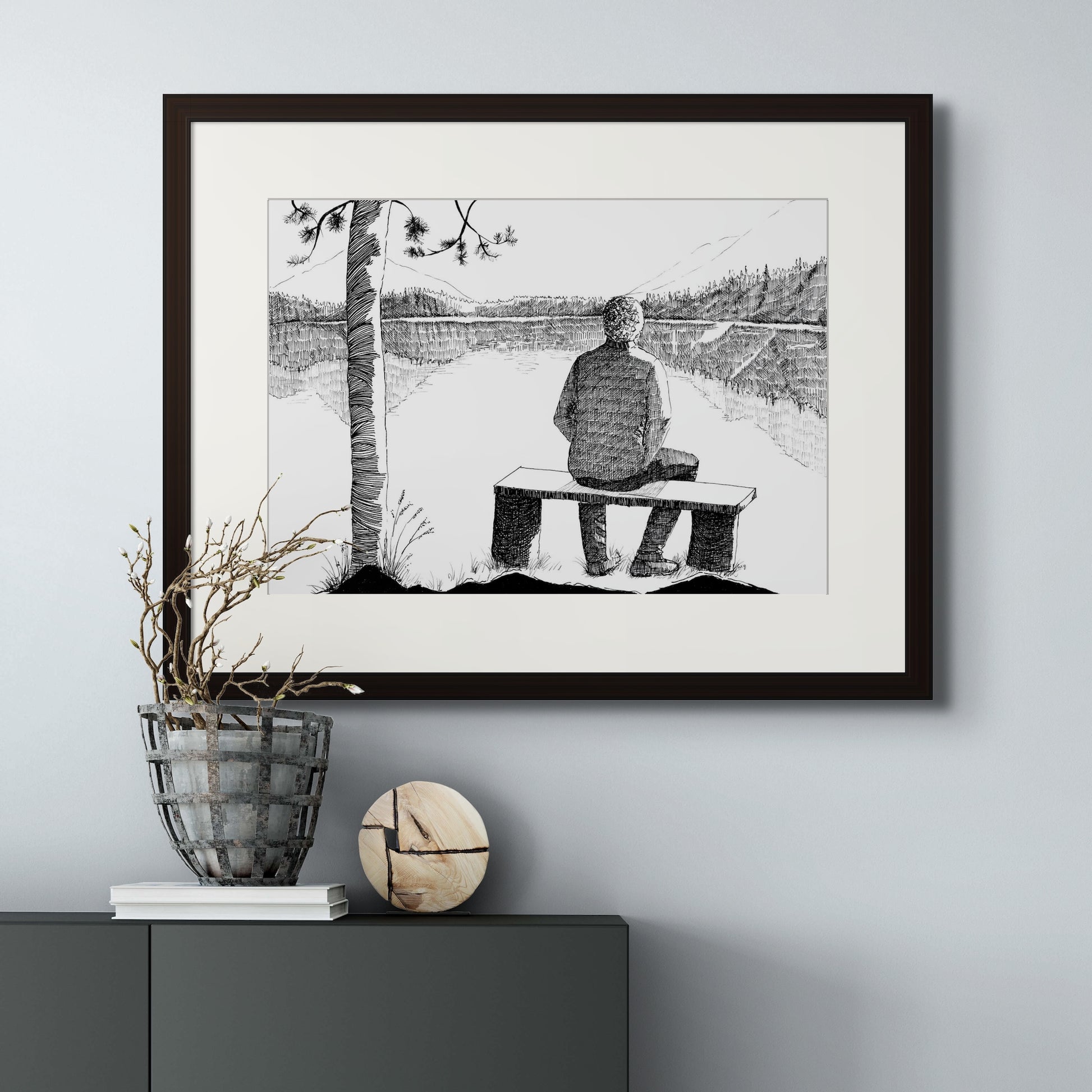 Image: line drawing landscape scenery of lake and mountain with a man seated on a bench. Giclée art print with black frame and cream colour mat.